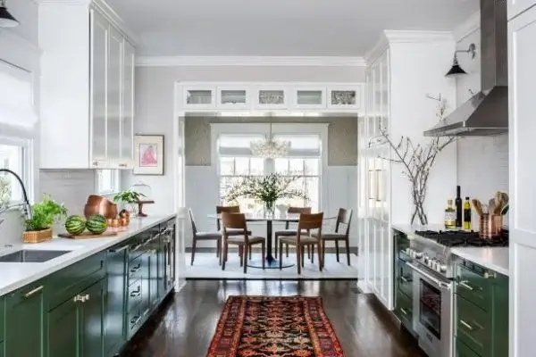 Traditional kitchen with white upper cabinets, dark green lower cabinets, stainless steel appliances, and a red patterned runner rug, leading to a bright dining area with wooden chairs.