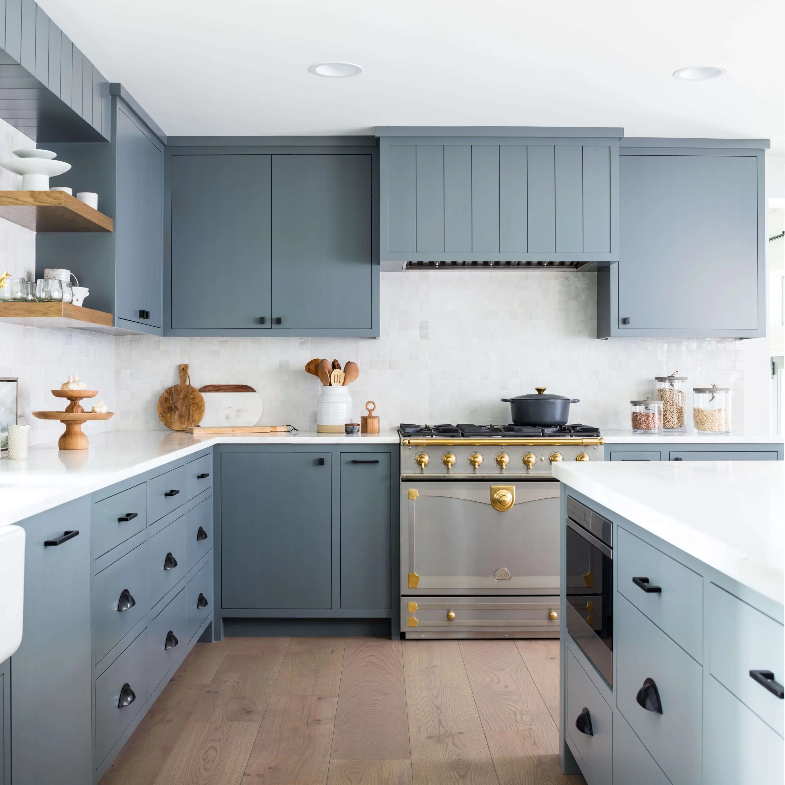 Modern kitchen with blue cabinets, a stainless steel and gold stove, white countertops, and open wooden shelves displaying kitchenware